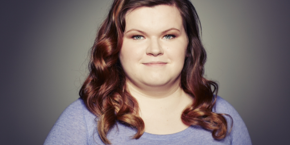 A head shot of Nikki. She is looking directly into the camera, smiling slightly with curly brown hair and a purple t shirt.